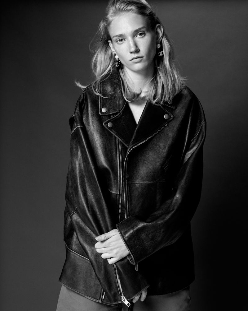 Black and white portrait of a young woman with light hair wearing a dark, oversized leather jacket against a grey background.