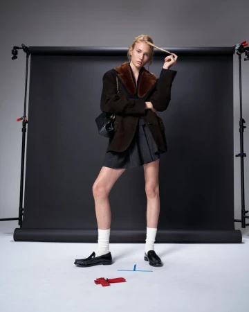 Model Tess Breeden wearing a brown coat and black loafers against a black backdrop