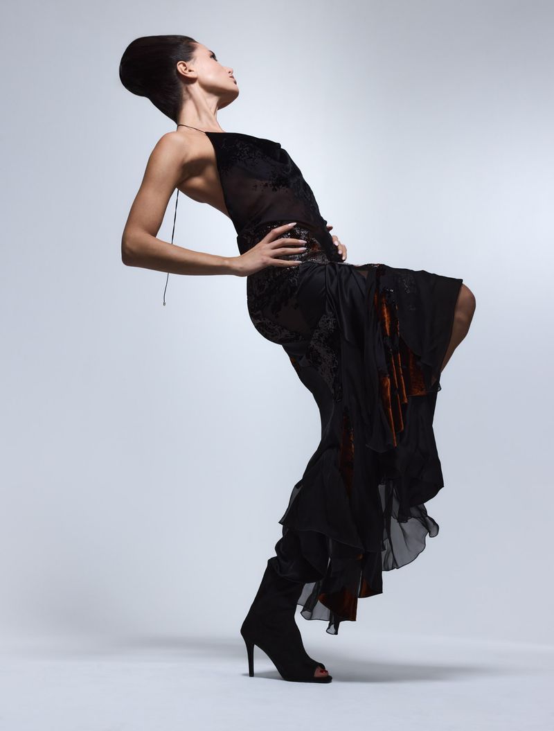 A stylish adult female poses in a long black dress and high-heeled boots against a pale background in a studio fashion shot.