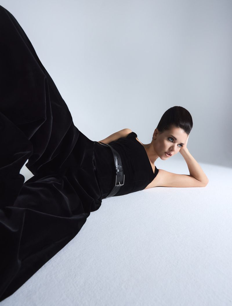 Elegant woman in black velvet jumpsuit with belt, laying on a light gray surface in a fashion photography studio setting.