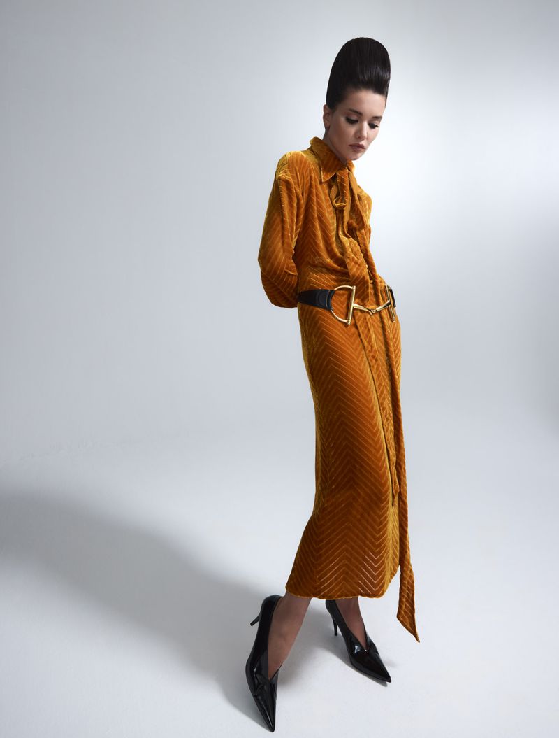 A woman in a gold, patterned dress with black heels and a black belt poses in a studio with a neutral background.