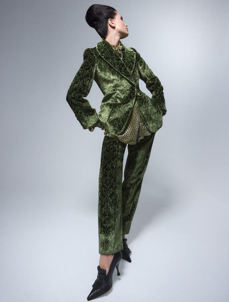 Elegant woman in green velvet suit poses against grey backdrop in studio shot. Hair is styled up. Black stiletto heels complete look.