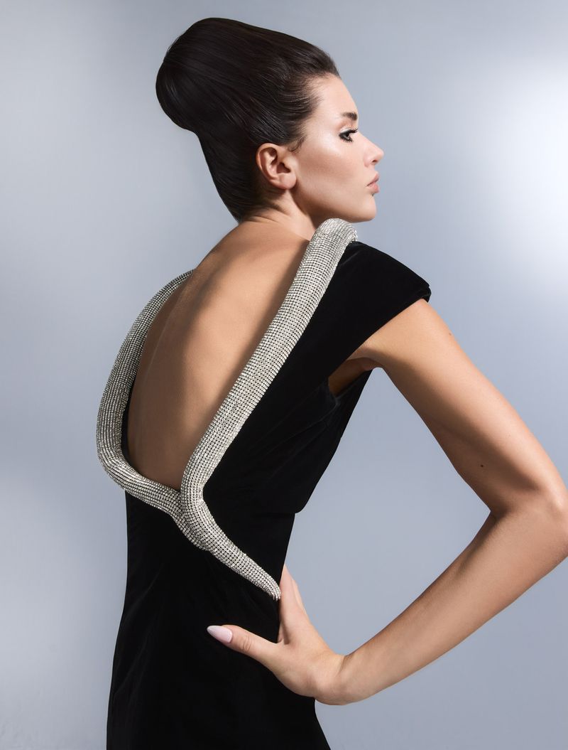 Woman with elegant updo poses in a black sleeveless dress with rhinestone straps, her hand on her hip, set against a gray backdrop.
