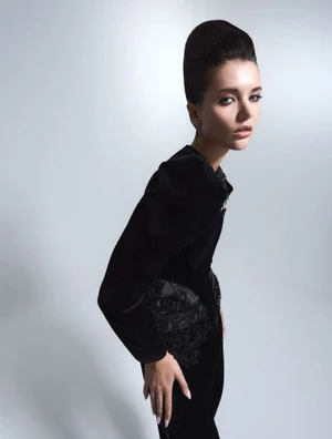 Woman with a retro bouffant hairstyle wearing a black velvet garment against a light grey studio background.