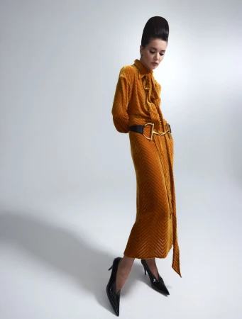 Model with high bouffant hairstyle wearing a mustard-yellow textured velvet dress and black heels against a pale studio backdrop.