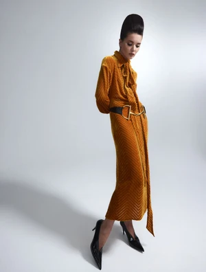 Model with high bouffant hairstyle wearing a mustard-yellow textured velvet dress and black heels against a pale studio backdrop.