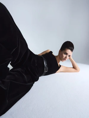 Model wearing a black velvet jumpsuit with a belted waist reclining on a pale studio floor.