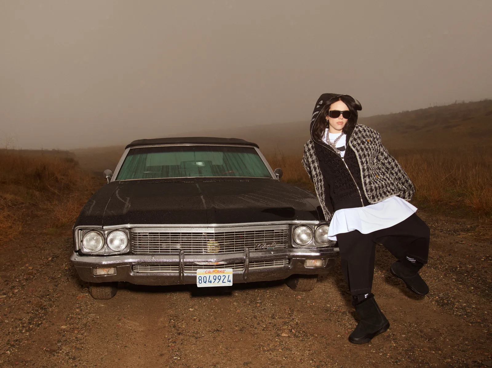 Woman poses by a classic black car on a dirt road in foggy weather, wearing a houndstooth jacket and sunglasses in an outdoor setting.