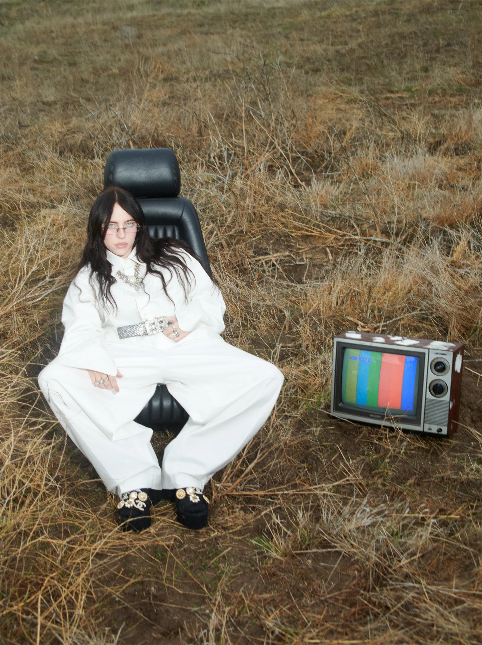 A young woman with dark hair sits in a black car seat in a grassy field, next to a retro television set with a color bar display.