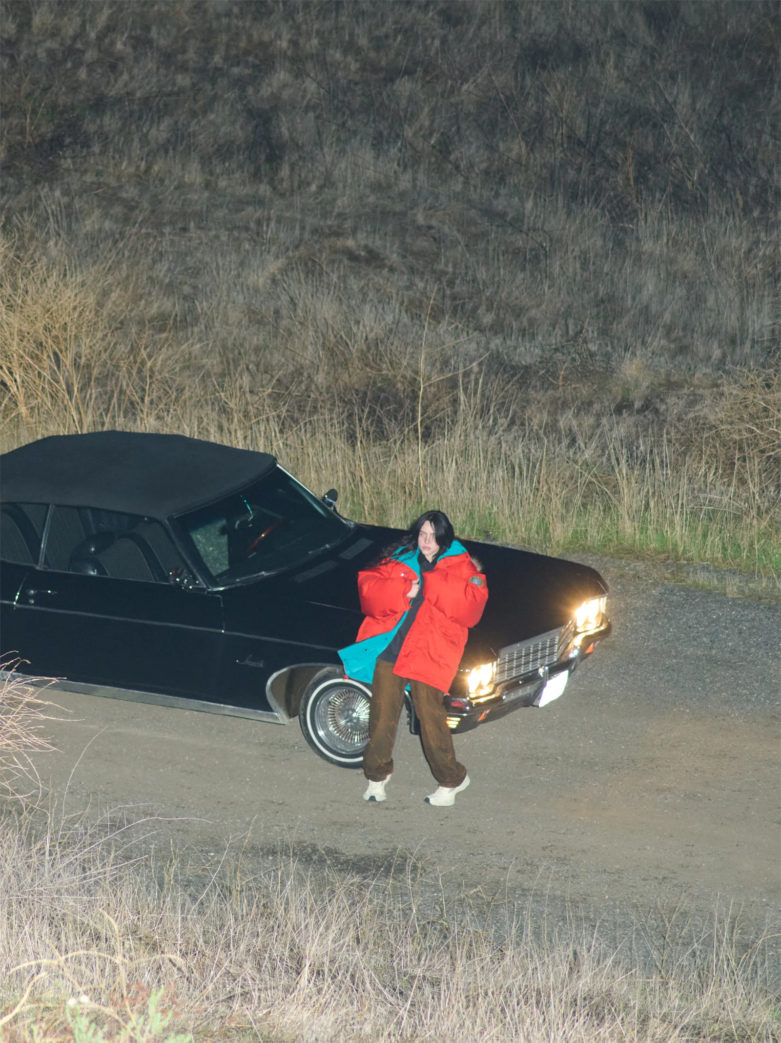 A person in an orange jacket poses on a dirt road next to a black convertible with its headlights on, set against a background of dried grass.