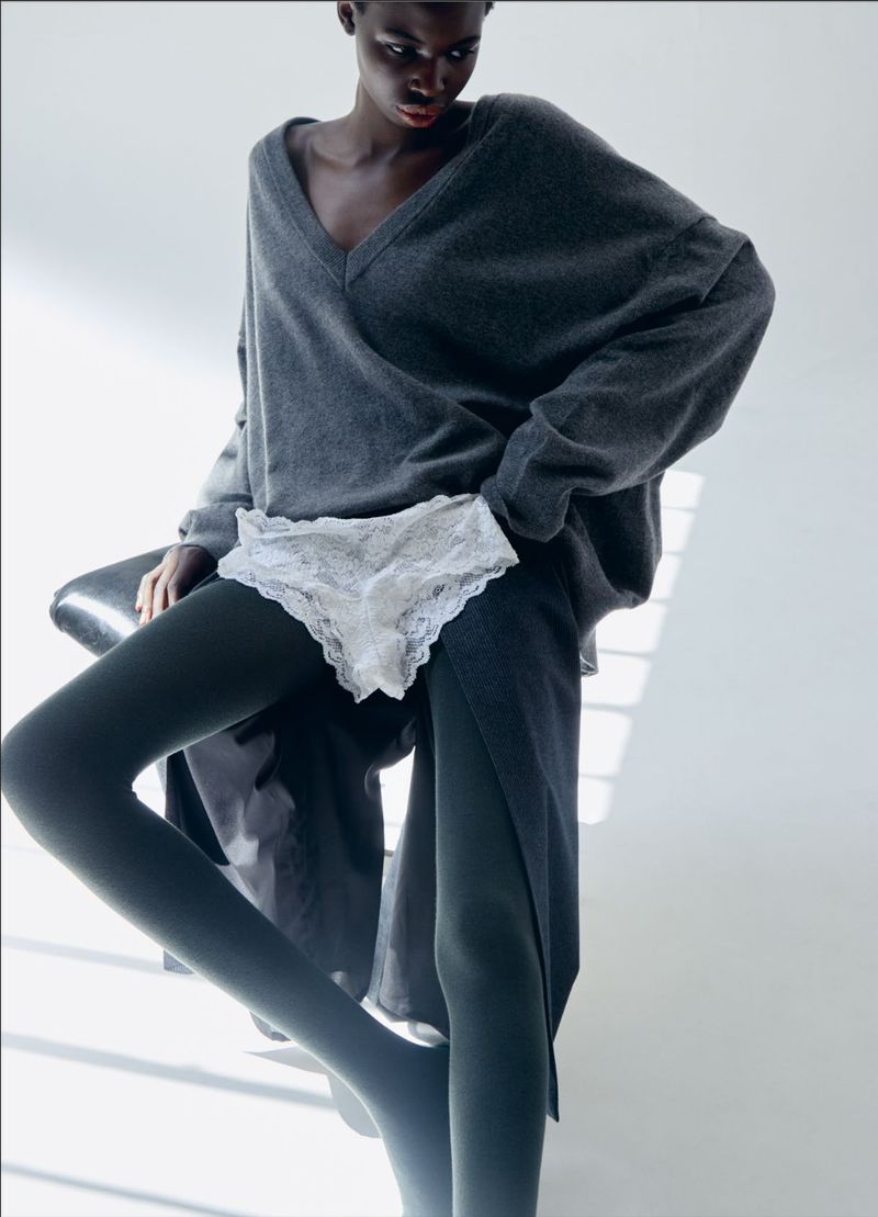 A dark-skinned woman poses indoors, showcasing a grey oversized sweater and black tights, seated on a chair, in a fashion editorial.