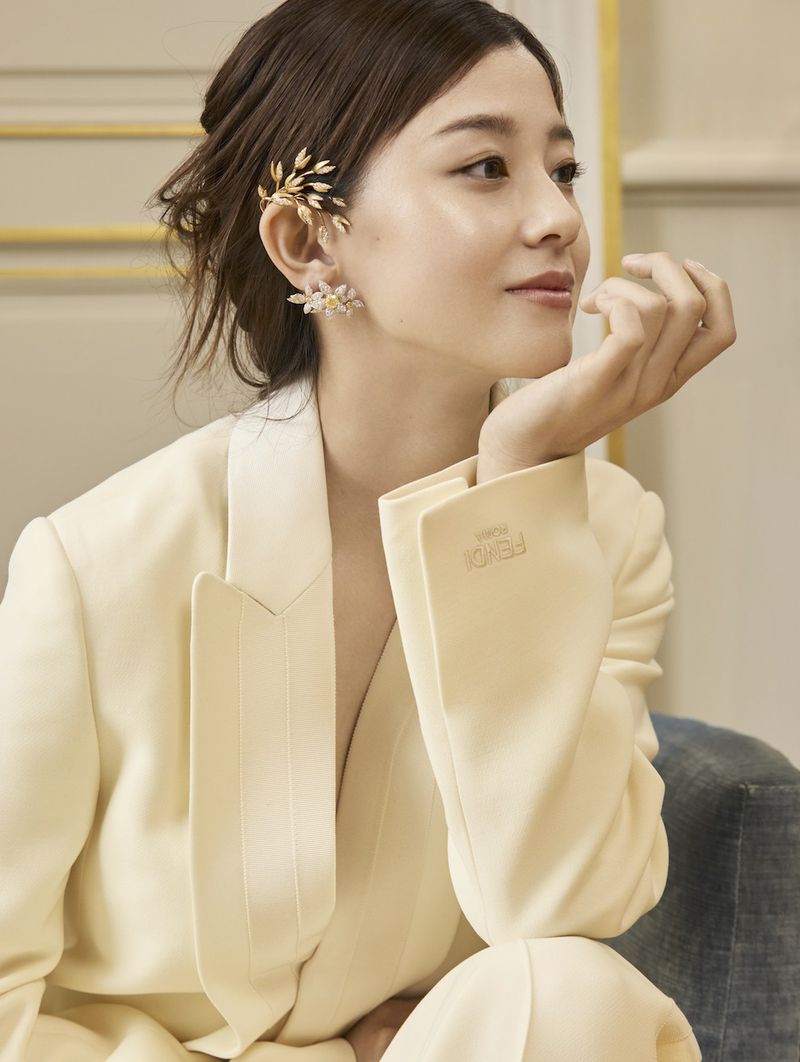 Woman wearing gold leaf ear cuffs and a cream suit jacket resting her chin on her hand in an indoor setting.