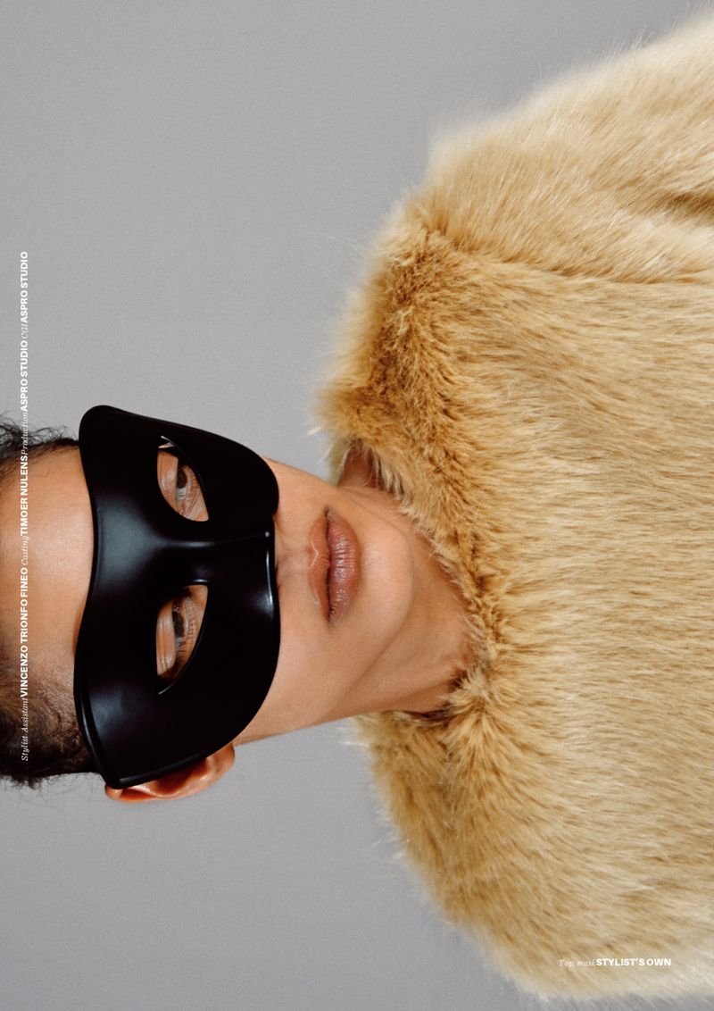 Portrait of a woman wearing a fur coat and a black mask against a grey background in a studio setting.