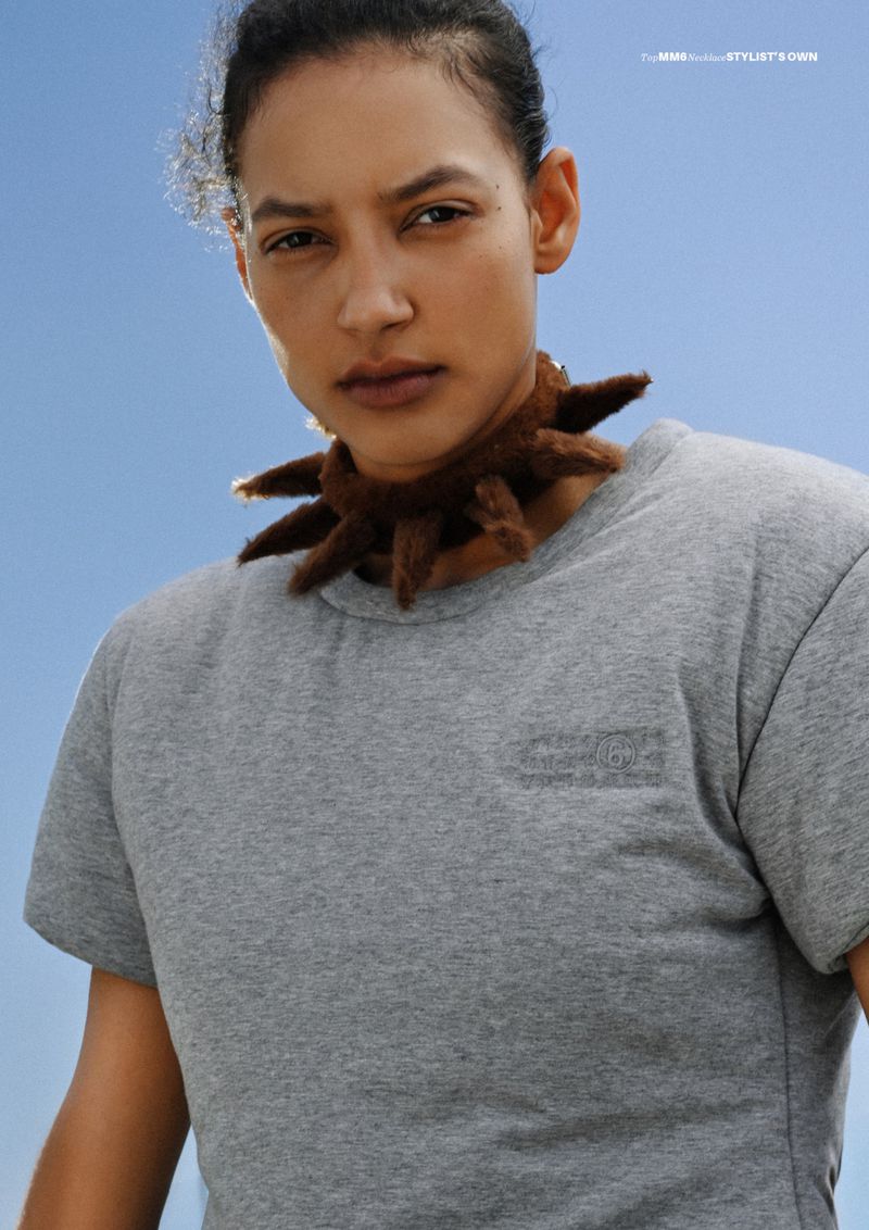 Model wearing a grey t-shirt and a spiky brown collar necklace against a clear blue sky background.