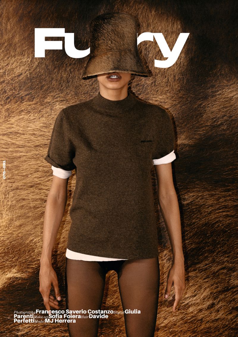 Fashion editorial shot with a full-length view of a woman wearing a brown sweater, tights, and a unique furry hat, set against a textured brown background.