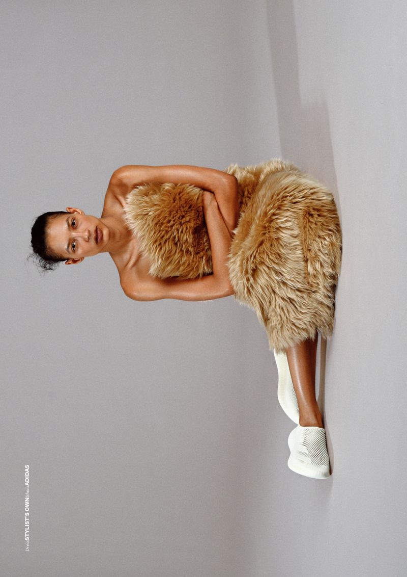 Model wearing fluffy fur dress and white sneakers seated against a neutral grey studio background