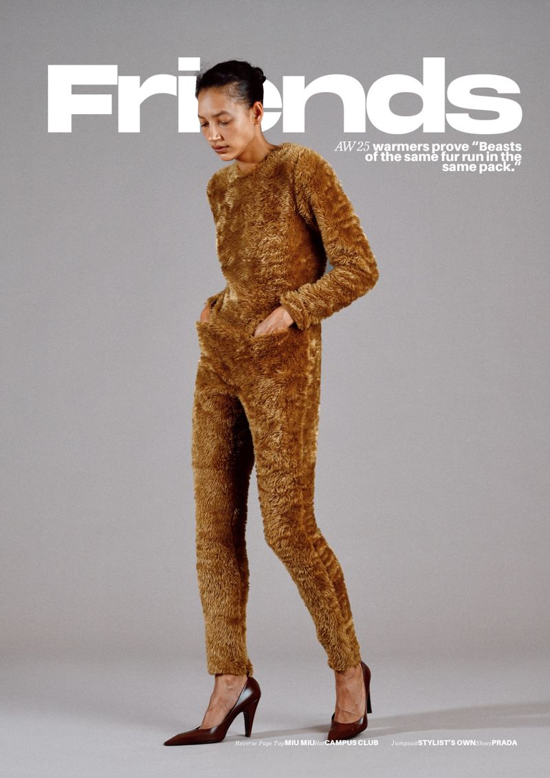 Editorial fashion shot of a slender adult woman in a furry, brown, long-sleeved jumpsuit, and matching heels, standing against a grey background.