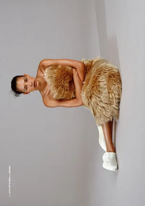 Model wearing fluffy fur dress and white sneakers seated against a neutral grey studio background
