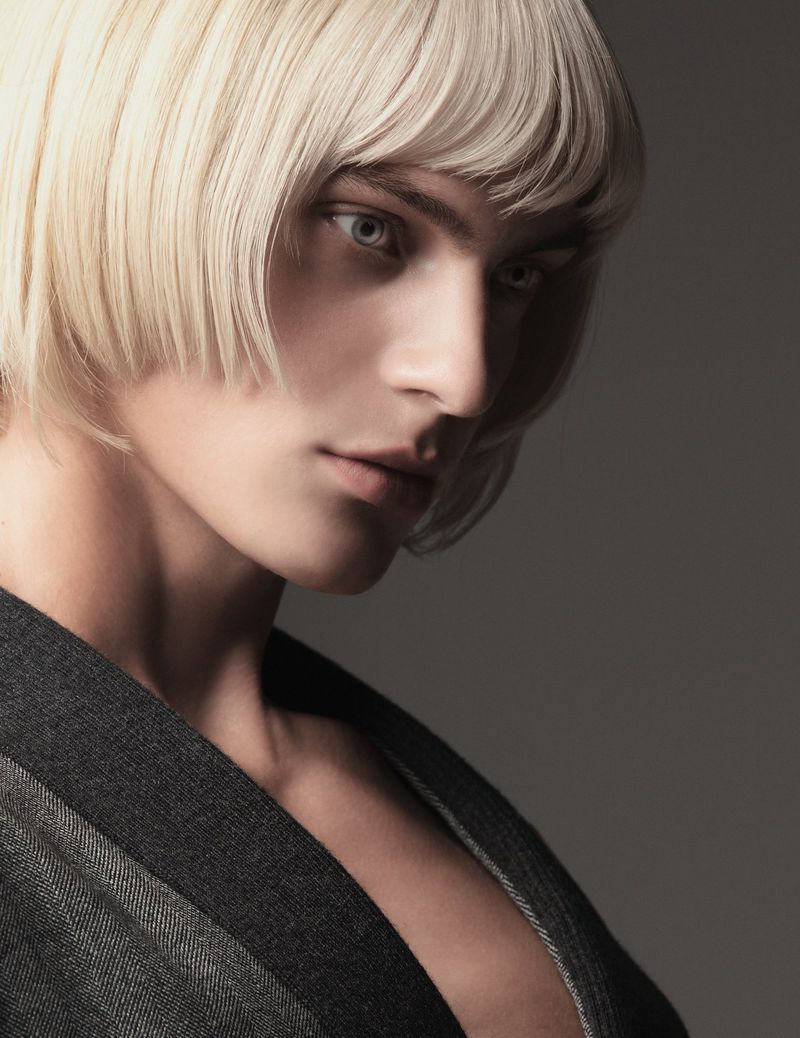 Striking close-up studio portrait of a young adult with short blonde hair and a grey striped jacket, against a neutral grey backdrop.