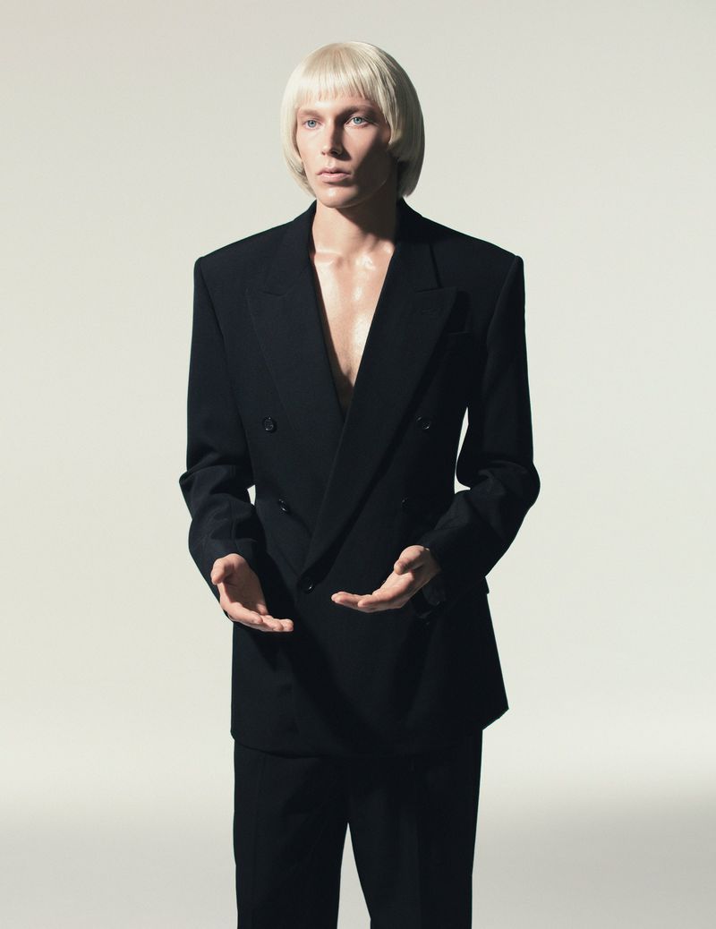 Elegant, fashion-forward portrait of a blonde male model in a black double-breasted suit, set against a neutral backdrop in a photo studio.