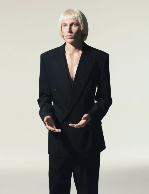 Elegant, fashion-forward portrait of a blonde male model in a black double-breasted suit, set against a neutral backdrop in a photo studio.
