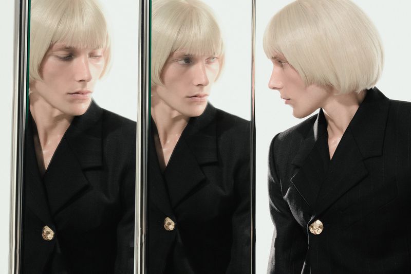 Person with blond bob and black jacket poses in front of three mirrors in a studio with white walls and ample lighting.