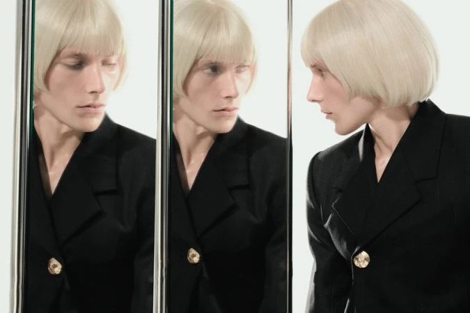 Person with blond bob and black jacket poses in front of three mirrors in a studio with white walls and ample lighting.
