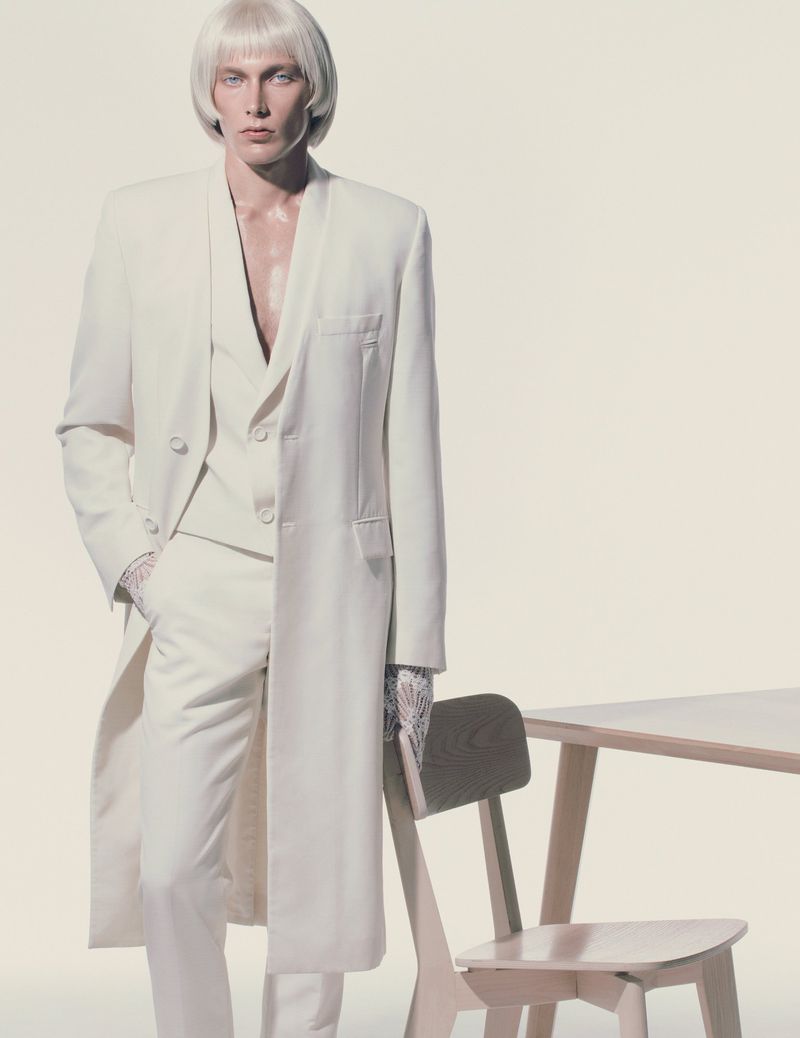 Fashion shot of a man with a platinum wig wearing a white suit and standing next to a table with a light background.