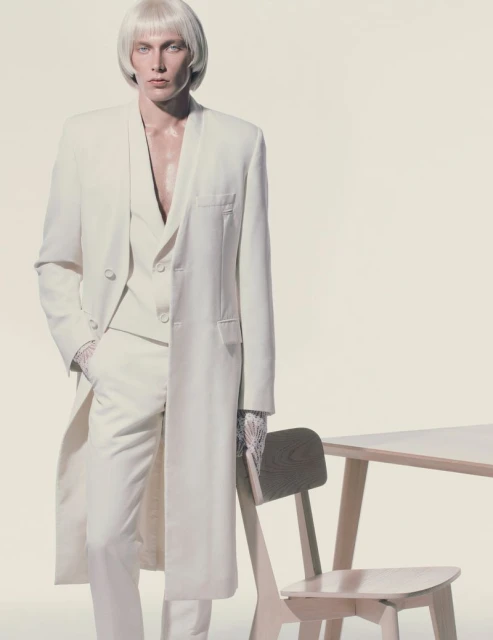 Fashion shot of a man with a platinum wig wearing a white suit and standing next to a table with a light background.