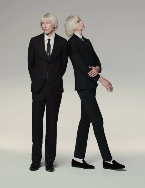 Two adult men with blonde hair wearing black suits and ties stand in a photo studio against a light gray background, striking a fashion pose.