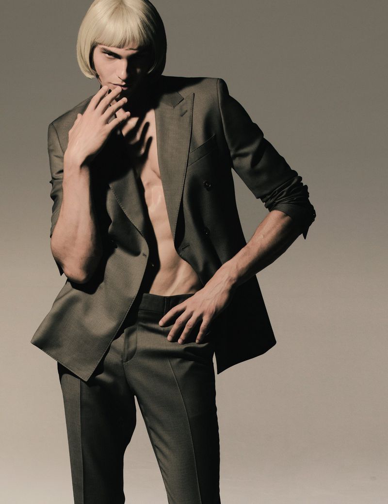 Man in a dark suit and a blonde wig posing in a studio with his hand near his mouth. Fashion shot of a male model.