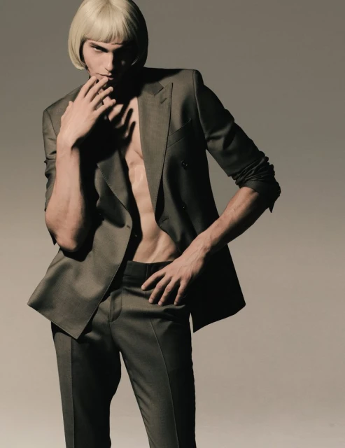 Man in a dark suit and a blonde wig posing in a studio with his hand near his mouth. Fashion shot of a male model.