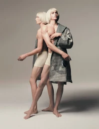Two male models in matching blonde bob wigs and beige briefs share a houndstooth coat against a grey studio backdrop.