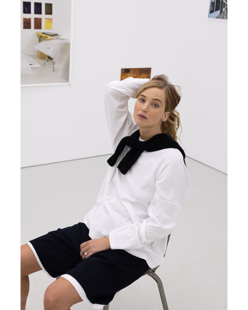 Woman in white long-sleeved shirt and dark shorts sits in a photo studio with a black sweater around her neck.