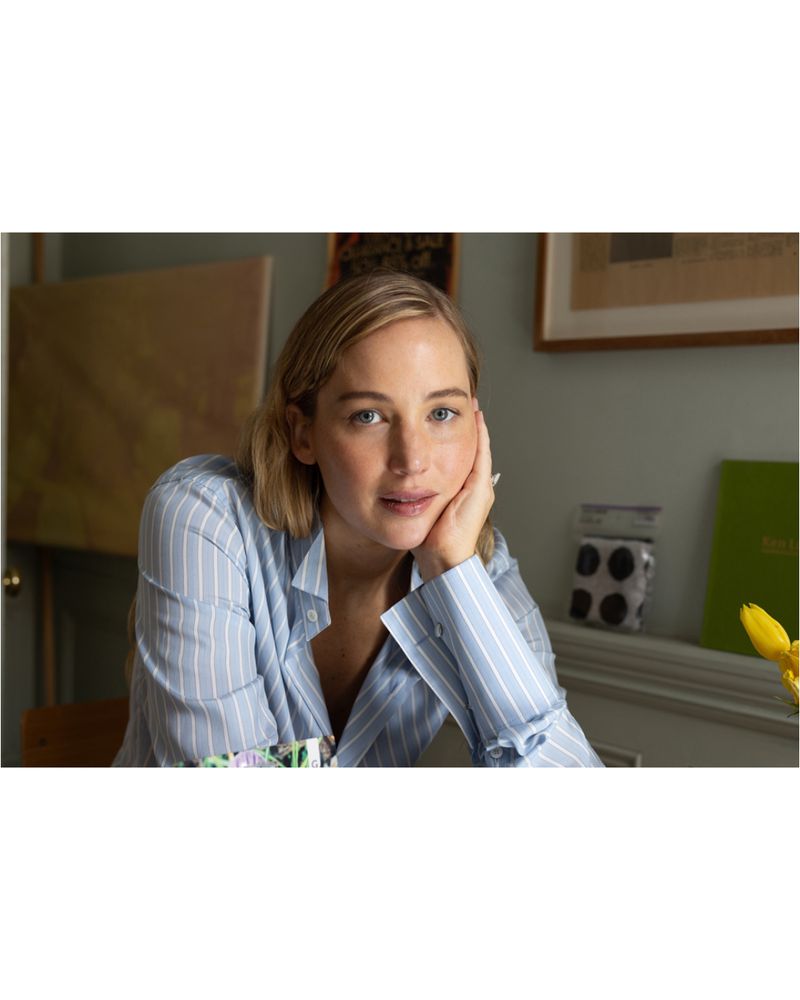 Jennifer Lawrence poses indoors wearing a blue striped shirt. Artwork and household items are visible in the background.