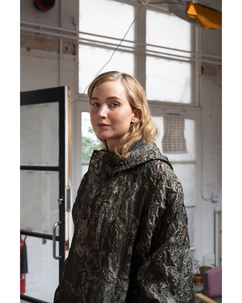 Portrait of a blonde woman in a textured, hooded poncho posing indoors with natural lighting and a neutral expression in a medium shot.