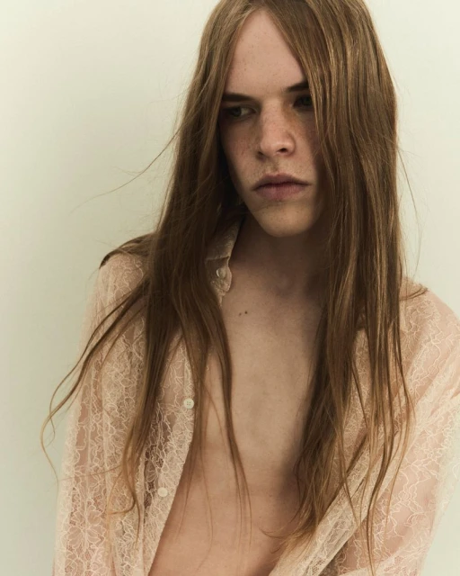 Portrait of a young adult with long brown hair and a light pink lace shirt, posing against a neutral background in soft, diffused light.