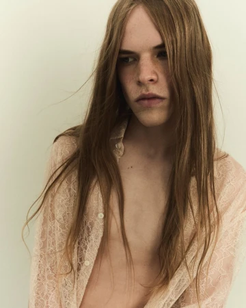 Portrait of a young adult with long brown hair and a light pink lace shirt, posing against a neutral background in soft, diffused light.