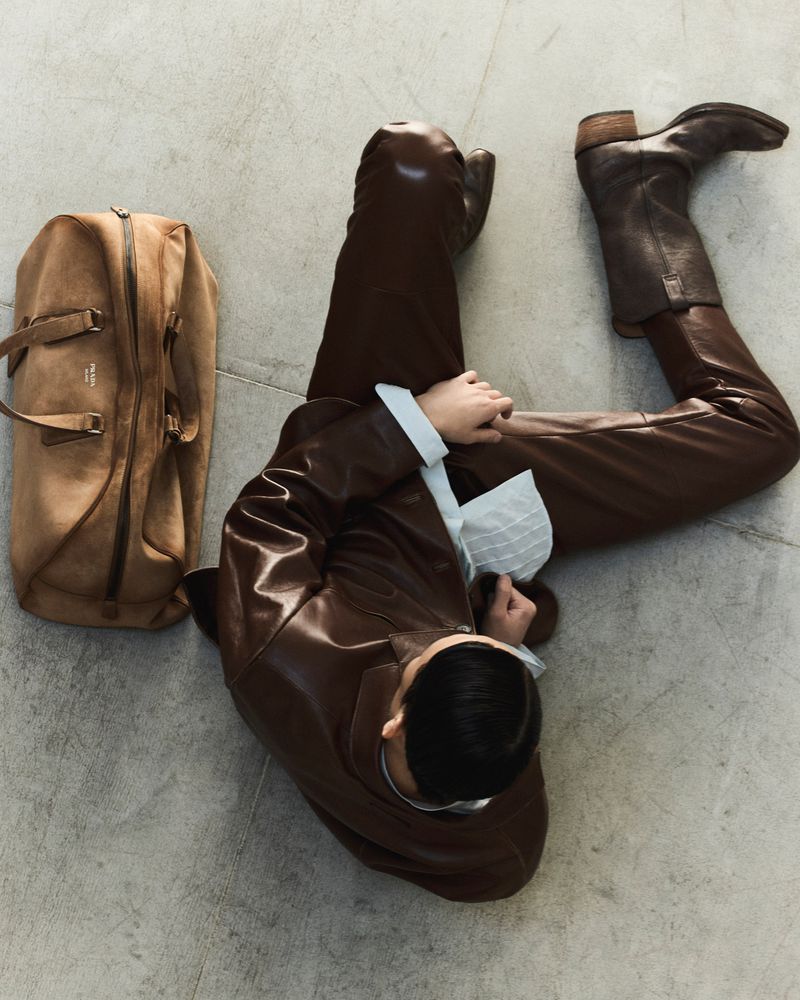 Overhead view of person in brown leather outfit and boots, sitting with tan duffel bag on a grey floor, high fashion.