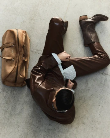 Overhead view of person in brown leather outfit and boots, sitting with tan duffel bag on a grey floor, high fashion.