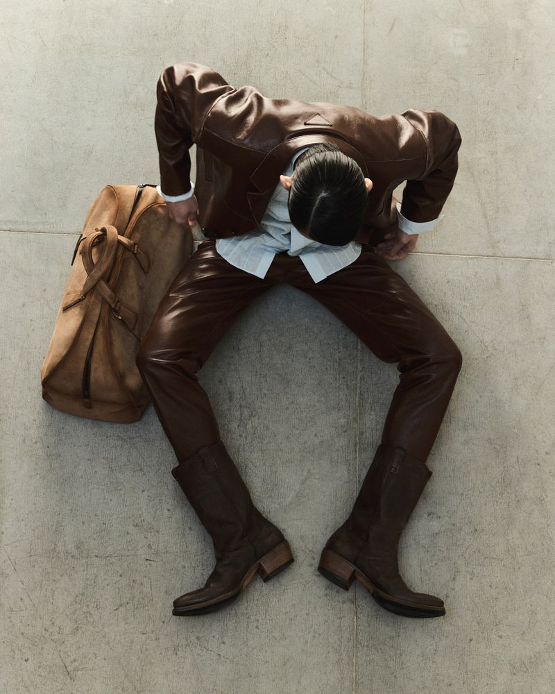 Overhead shot of a man wearing brown leather suit and boots standing on a gray floor with a brown duffel bag on the floor.