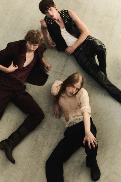 Three men laying on a concrete floor, wearing leather, lace and velvet clothing in a dimly lit indoor environment. Fashion editorial.