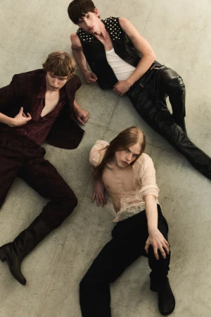 Three men laying on a concrete floor, wearing leather, lace and velvet clothing in a dimly lit indoor environment. Fashion editorial.