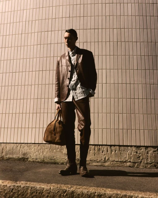 A slim, fair-skinned male model poses in a brown leather jacket, patterned shirt, and leather trousers on a city street with a brown duffel bag.