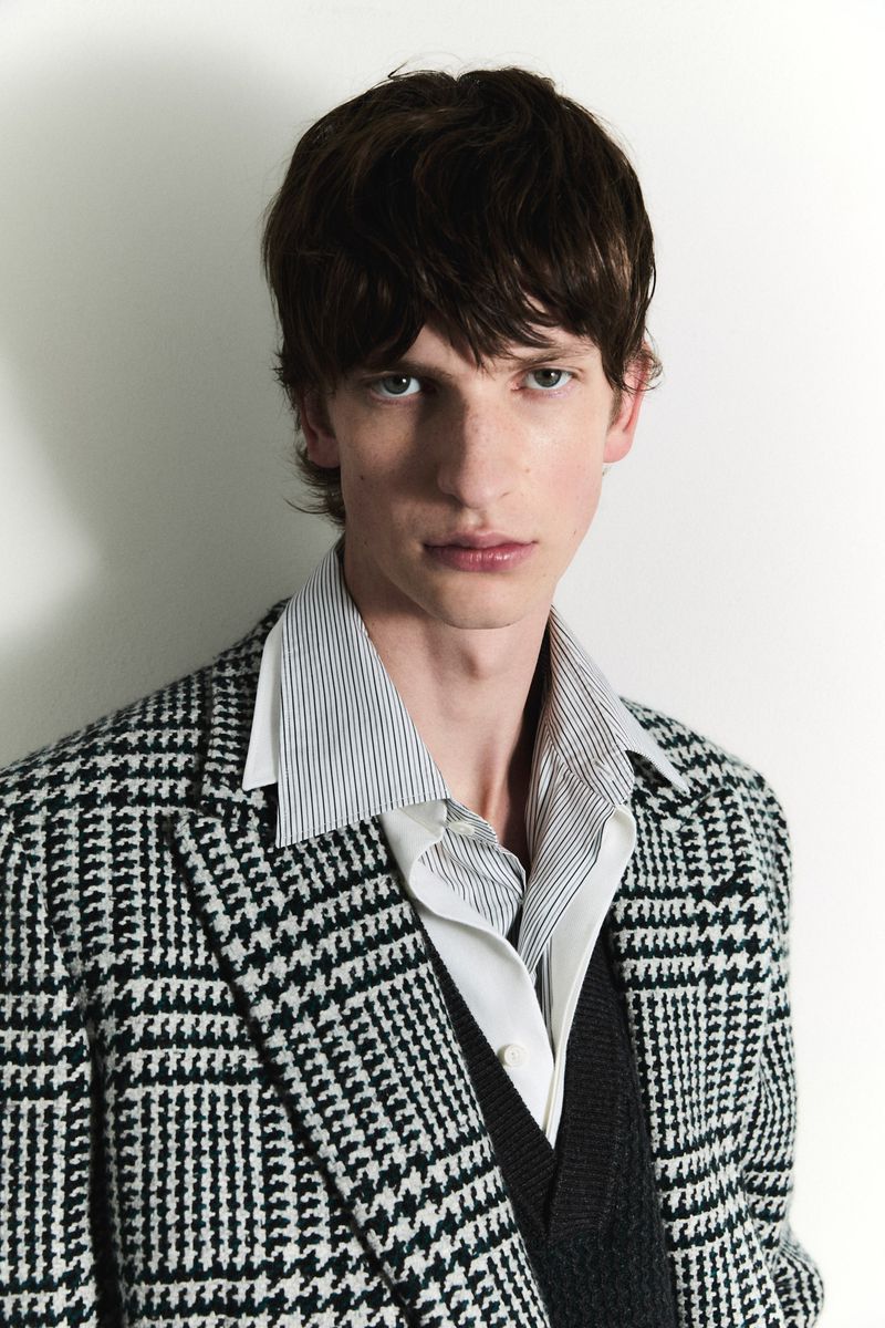 A portrait of a light-skinned male model with brown hair wearing a black and white patterned blazer, a striped shirt, and a v-neck sweater.