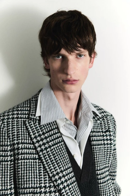 A portrait of a light-skinned male model with brown hair wearing a black and white patterned blazer, a striped shirt, and a v-neck sweater.