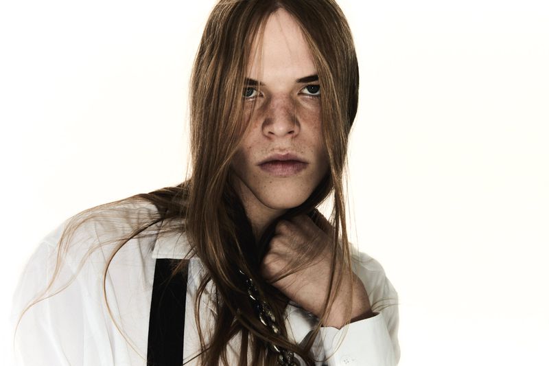 Portrait of a young adult with long brown hair, freckles, wearing a white shirt against a bright white background in a photo studio.