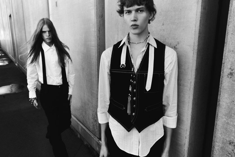 Two androgynous fashion models in black and white pose in an urban setting. One model wears a vest, and the other has suspenders.