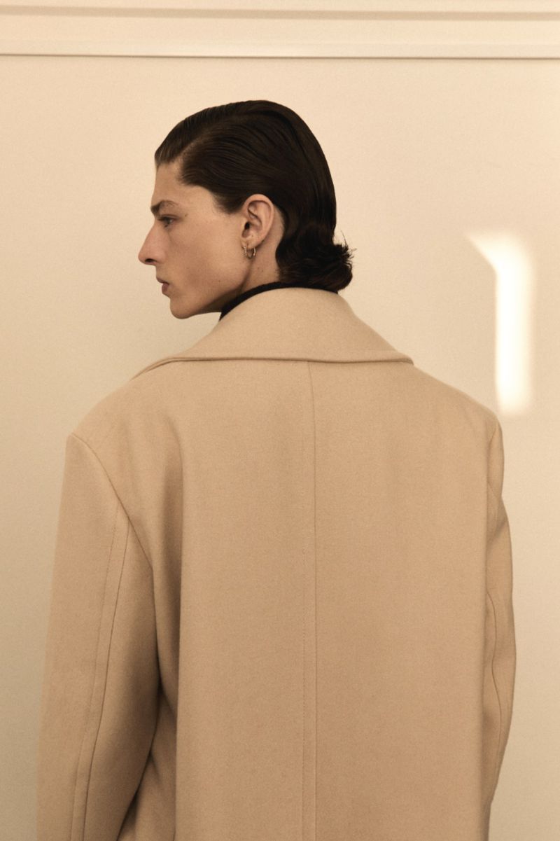 Side profile of a male adult wearing a beige coat against a white wall.  Dark hair is neatly styled in a vintage manner. Indoor fashion shot.