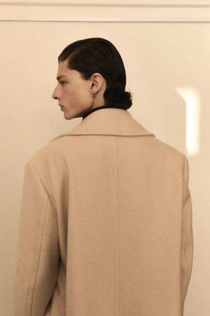 Side profile of a male adult wearing a beige coat against a white wall.  Dark hair is neatly styled in a vintage manner. Indoor fashion shot.